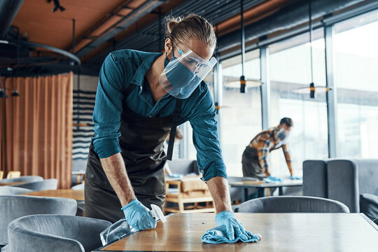 Young male waiters in protective workwear cleaning tables in restaurant