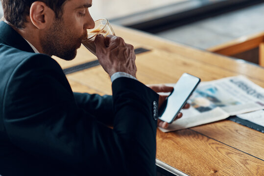 Handsome young businessman in full suit using smart phone and drinking coffee while sitting in the restaurant