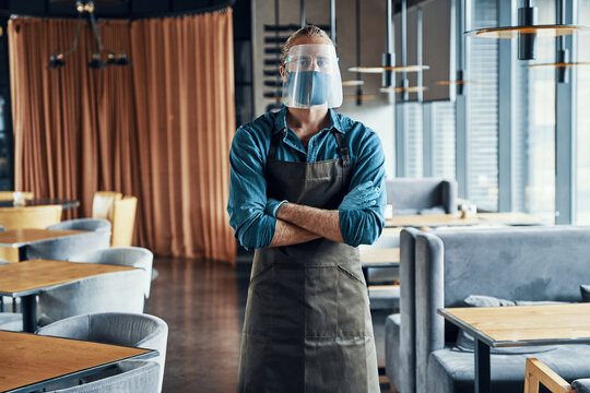 Confident male waiter in protective face shield keeping arms crossed