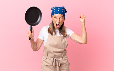 pretty middle age woman shouting aggressively with an angry expression and holding a pan. chef...