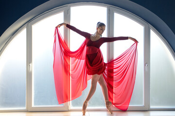 Beautiful young focused ballerina in elegant long red ballet dress in butterfly pose stands in front of big round shaped window at ballet studio