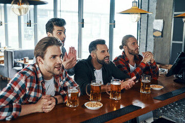 Cheering young men in casual clothing drinking beer and watching sport game while sitting in the pub