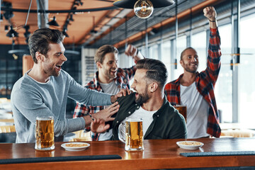 Cheering young men in casual clothing drinking beer and watching sport game while sitting in the pub