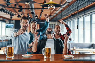 Excited young men in casual clothing drinking beer and watching sport game while sitting in the pub