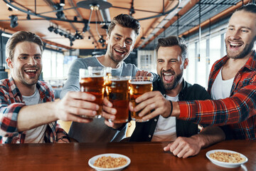 Happy young men in casual clothing toasting each other with beer and laughing while sitting in the pub