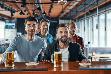 Excited young men in casual clothing drinking beer and watching sport game while sitting in the pub