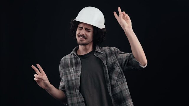 The cheerful, charismatic engineer brings his fingers in peace and dances modestly and happily. Isolated on black background. The concept of life. People's emotions. 4k portrait