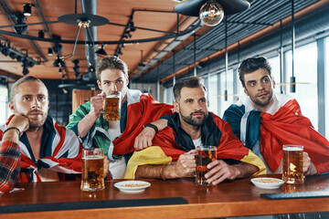 Young men covered in international flags drinking beer and watching sport game while sitting in the pub