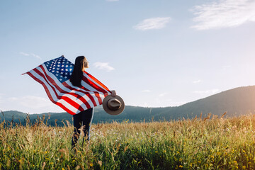 Beautiful young american brunette woman in hat standing on grass of background mountains in wind independent and young, holds proudly usa flag high in air, demonstrates patriotism and freedom - Powered by Adobe