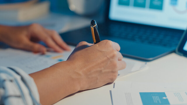 Close-up young asia lady freelance focus on laptop write worksheet finance graph account chart market plan in laptop and tablet at house. Girl student learn online, Work from home night concept.