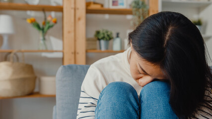 Thoughtful Asia lady sit embracing knees at sofa in living room at house look outside with feel lonely, Sad depressed teenager spend time alone stay at home, Social distance, Coronavirus Quarantine.