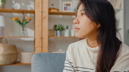 Thoughtful Asia lady sit embracing knees at sofa in living room at house look outside with feel lonely, Sad depressed teenager spend time alone stay at home, Social distance, Coronavirus Quarantine.