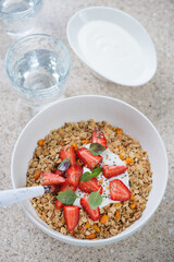 Bowl of granola topped with yogurt and fresh strawberries, vertical shot on a beige marble background, elevated view