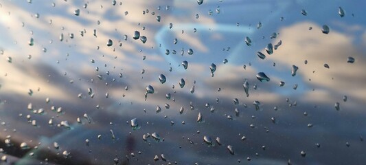 Reflection of the sky from wet glass. There are several large drops of water on the car glass, which is wet after rain. The blue sky and white-gray clouds are reflected from the glass.
