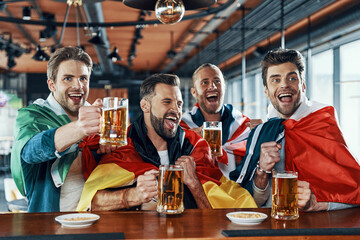 Happy young men covered in international flags drinking beer and watching sport game while sitting in the pub