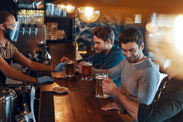 Smiling young men in casual clothing drinking beer and bonding together while sitting in the pub