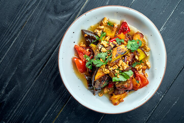 Appetizing, dietary salad of baked vegetables (tomatoes, peppers, eggplants) with sauce and nuts in a white plate on a dark wooden background. Vegetarian food. Selective focus