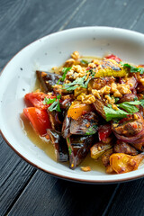 Appetizing, dietary salad of baked vegetables (tomatoes, peppers, eggplants) with sauce and nuts in a white plate on a dark wooden background. Vegetarian food. Selective focus
