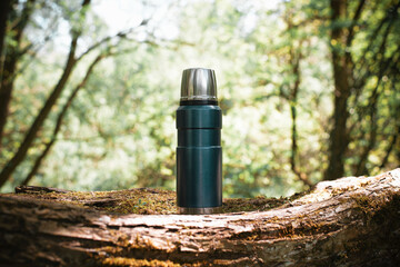 Thermos standing on a tree outdoors, blurred backdrop forest sunny summer day. Hiking flask with a hot drink in nature. Camping concept. - obrazy, fototapety, plakaty