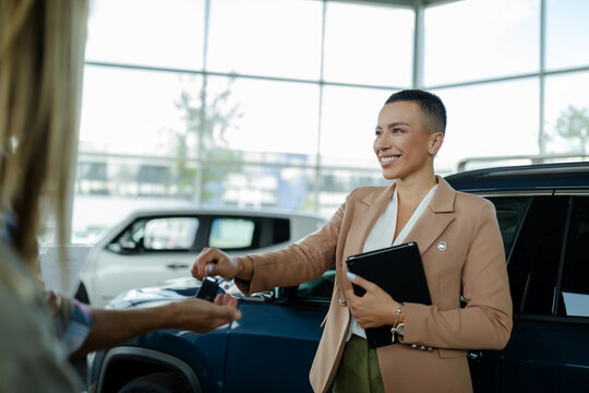 Beautiful young saleswoman in car dealership giving keys to clients.
