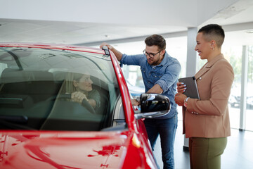 Young smiling saleswoman is holding a tablet and showing new car to couple in showroom.
