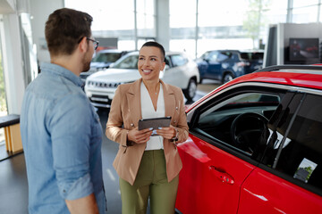 Handsome man with car dealer in auto show or salon. Saleswoman is holding a tablet in hands.
