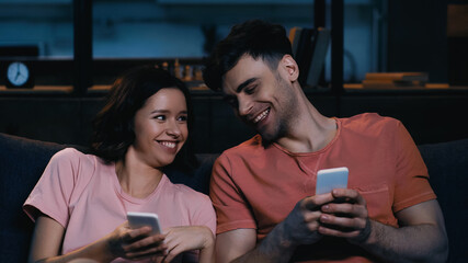 cheerful man and woman holding cellphones and smiling in modern living room.