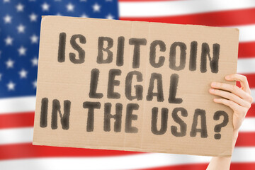 The phrase " Is Bitcoin legal in the USA? " on a banner in men's hand with blurred American flag on the background. Currency. Legislation. Financial system. Economy. Savings. Money