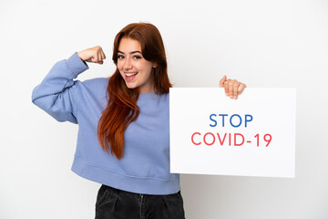 Young redhead woman isolated on white background holding a placard with text Stop Covid 19 and doing strong gesture