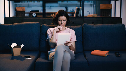 brunette woman using digital tablet while sitting on sofa near smartphone, book and carton box with food.