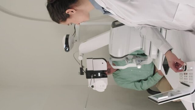 Vertical medium shot of young female optometrist using ophthalmic eye chart projector while doing eyesight test for senior man sitting in chair at appointment