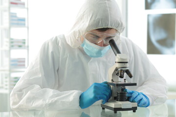 Fototapeta na wymiar Doctor in a protective medical suit wearing a mask wearing blue gloves in a laboratory looks through a microscope