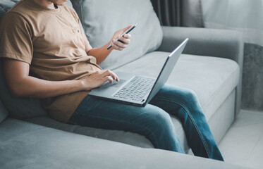 Naklejka premium young man typing on mobile phone while sitting on a sofa at home with laptop computer. Man using his smartphone for online banking.work from home concept.