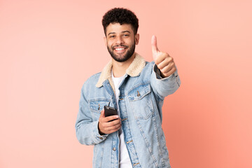 Young Moroccan man using mobile phone isolated on pink background with thumbs up because something...