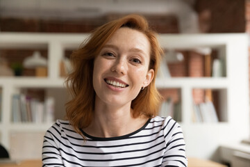 Head shot smiling attractive millennial red-haired businesswoman entrepreneur looking at camera, holding negotiations webcam video call remote meeting, enjoying distant communication in modern office.