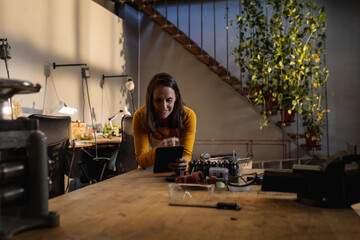 Caucasian female jeweller wearing apron, leaning on desk in workshop, using tablet, smiling