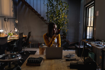 Caucasian female jeweller wearing apron, sitting at desk in workshop, using smartphone and laptop