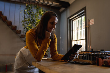 Caucasian female jeweller wearing apron, leaning on desk in workshop, using tablet, smiling