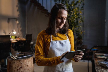 Focused caucasian female jeweller wearing apron, standing in workshop, using tablet