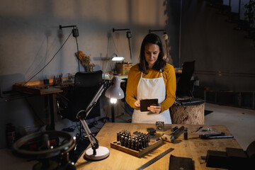 Focused caucasian female jeweller wearing apron standing at desk in workshop, using tablet