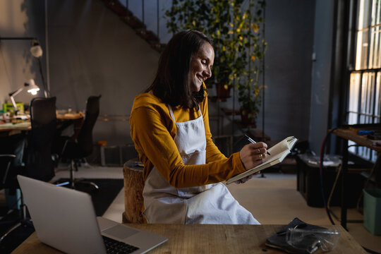 Smiling caucasian female jeweller wearing apron, sitting at desk, using smartphone, making notes