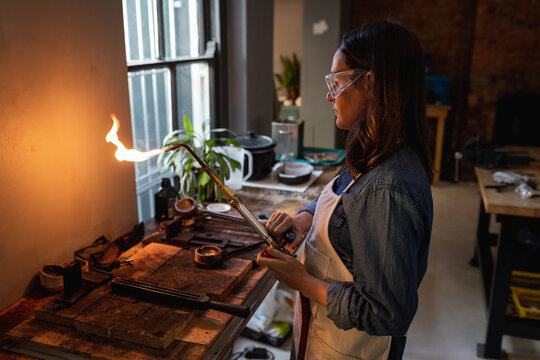 Caucasian female jeweller wearing apron and glasses, using gas burner, melting metal for jewelry