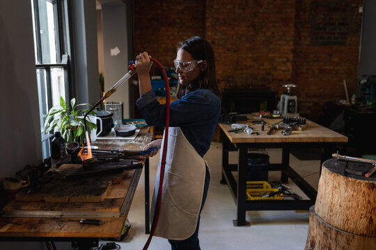 Caucasian female jeweller wearing apron and glasses, using gas burner, melting metal for jewelry - Powered by Adobe