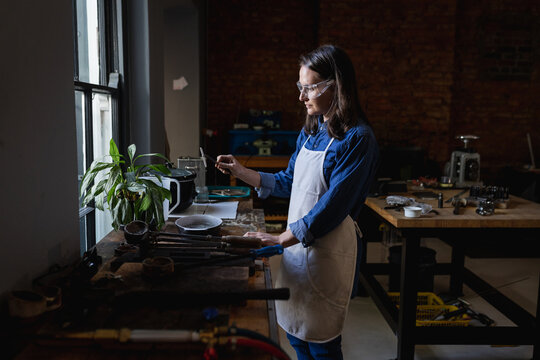 Caucasian female jeweller wearing apron and glasses, preparing materials for jewelry