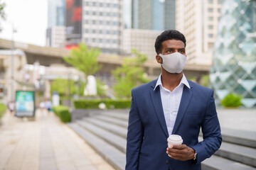 Portrait of African businessman wearing face mask outdoors in city and holding take away coffee cup