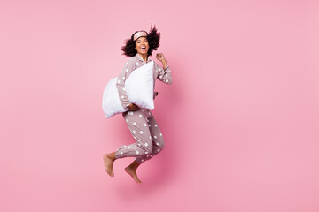 Photo of funky lucky wavy dark skin lady dotted sleepwear jumping high holding white pillow rising fist isolated pink color background