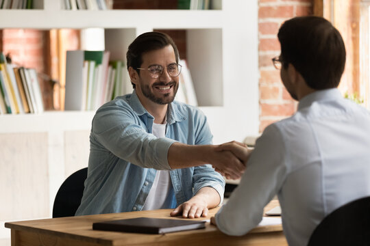 Smiling skilled handsome young male applicant in eyewear shaking hands with boss leader, accepting proposal or job offer, making good first impression on employer at meeting in modern office room.