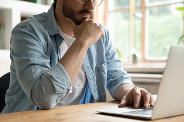 Obraz premium Cropped image thoughtful young man working on computer, sitting at table. Pensive millennial male manager worker employee analyzing electronic documents, considering problem solution, communicating.