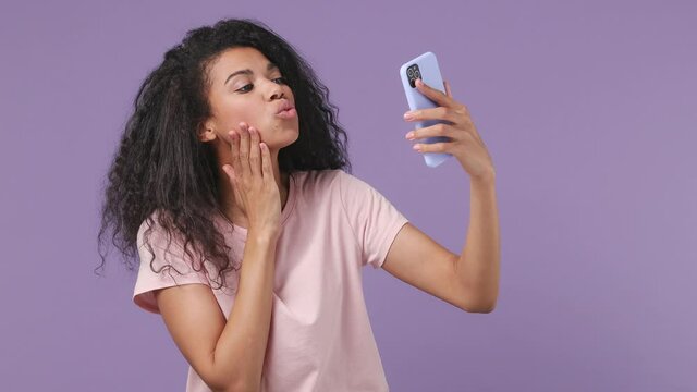 Young african woman 20s years old wears pink T-shirt doing selfie shot on mobile phone post photo on social network chatting blogging isolated on pastel violet purple color background studio portrait