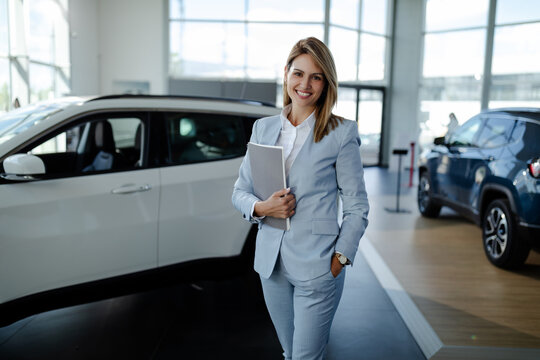 Female salesperson at a car showroom, holding a notes and smiling at the camera.